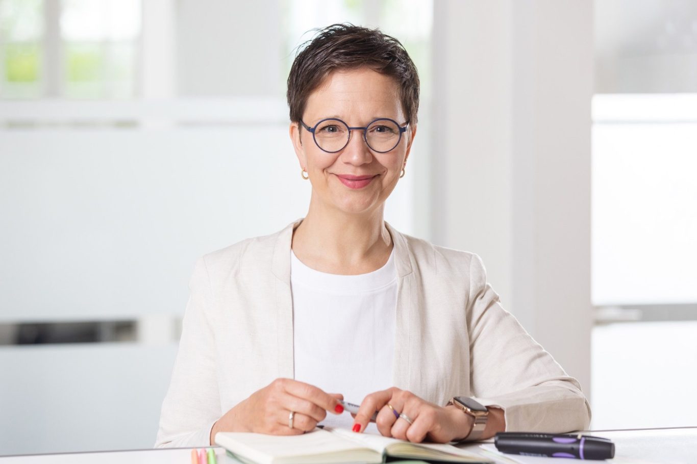 Anika Franke Das Bild zeigt Anika Franke, wie sie mit Kalender, Stift am Empfangstresen im Büro steht und in die Kamera lächelt.
