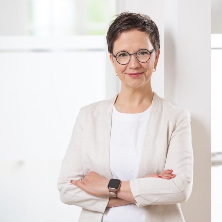 Das Bild zeigt Anika Franke vor einem Pfeiler stehend mit verschränkten Armen, wie sie in die Kamera lächelt. 