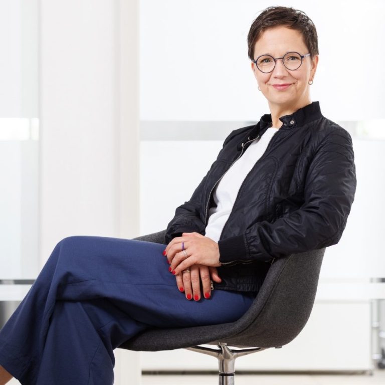 Anika Franke Das Bild zeigt Anika Franke, wie sie entspannt zurückgelehnt auf einem Stuhl sitzt und in die Kamera lächelt.